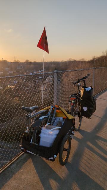Ein Trekkingrad mit Lastenanhänger steht auf der Erzbahn schwinge. In dem Anhänger steht ein Brompton-Faltrad und liegen mehrere Plastikcontainer. Am Gepäckträger des Rads hängt außerdem eine vollbepackte Ortliebtasche.