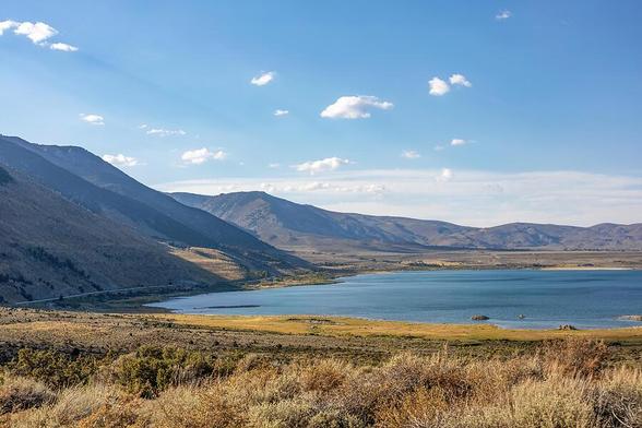 A scenic view of Mono Lake in California, surrounded by arid desert and mountainous terrain. The blue waters contrast with the dry vegetation and dramatic sky, creating a stunning natural scene.