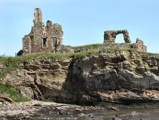 The crumbling and precarious clifftop ruins of Newark Castle. The foreground of the image has a bay with the sea to the right at quite low tide. There are cliffs in the middle ground beyond the bay with stone ruins on top of them. The sky is blue and the scene is in sunshine.