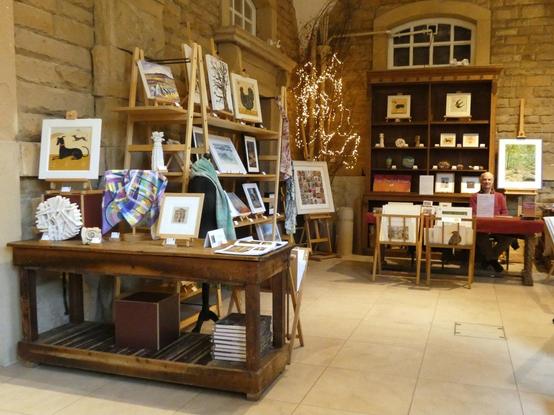 Inside a stone building old oak tables and a tall dresser display framed artwork, ceramics and textiles.