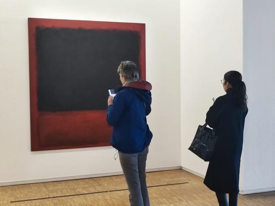 The first image of a series of three images in a row where we see two women taking turns to take a photo of a Mark Rothko artwork in Paris, Centre Pompidou (black and red rectangles on bloody red). A woman takes a photo while the other one waits her turn.