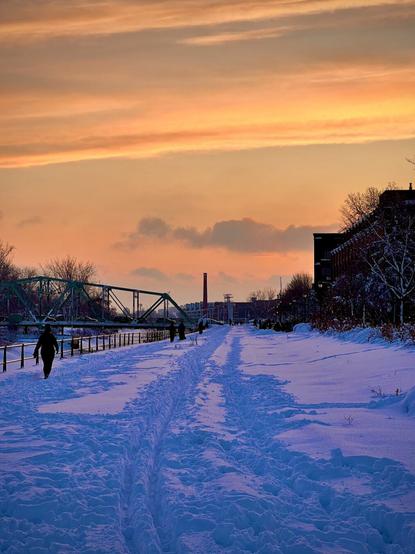 The scene depicts a snowy pathway along a river, with footprints and tire tracks visible in the snow. People are walking in the distance, and a bridge can be seen crossing the river. The sky is painted in warm hues of orange and pink, suggesting a sunset, while buildings and trees line the pathway, creating a serene winter atmosphere.