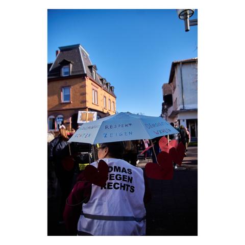 Foto: Eine ältere Frau ist zu sehen, mit einem Regenschirm auf dem steht  »Respekt zeigen« und sie trägt eine weiße Warnweste mit der Aufschrift »Omas gegen Rechts«