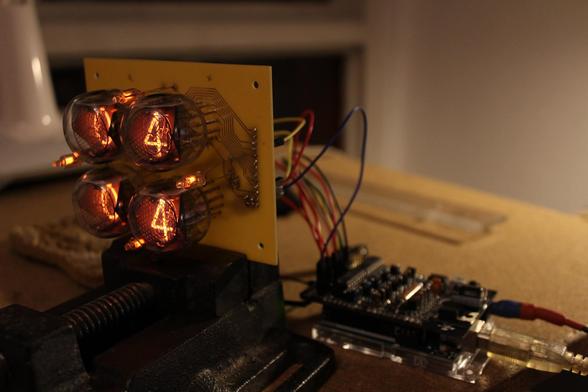A picture showing the mounting PCB of the nixie tubes in a vise, with the tubes displaying some digits. The Arduino control board and the cablework is visible on the right.