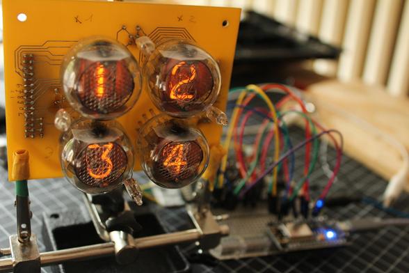 A picture showing the mounting PCB of the nixie tubes in a vise, with the tubes displaying some digits. An ESP32 control board and a breadboard with some cablework is visible in the background