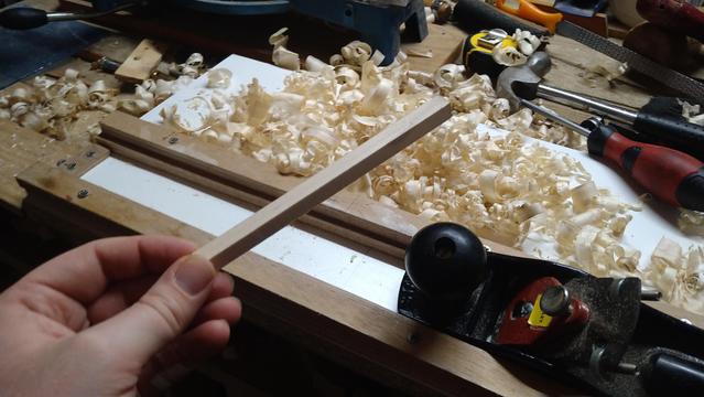 A hand holding a square profiled wooden stick, with a handplane, some wood shavings and other handtools in the background