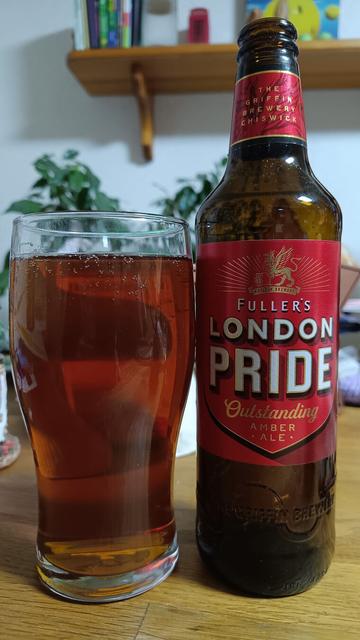 Auf einem Holztisch steht ein mit Bier gefülltes Glas und daneben eine Bierflasche mit Etikett auf dem auf roten Untergrund steht:
Fullers London Pride Outstanding Amber Ale
