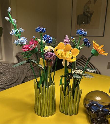 Two vibrant, LEGO flower arrangements in green, grass-like vases, featuring various colorful blooms. A transparent skull decoration is visible next to the flowers on a yellow table.
