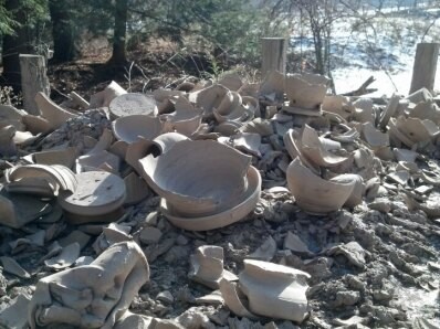 Broken, unfired, pottery and clumps of hardened clay. The sun is shining from the right, casting shadows on the broken bit and pieces. In the background are some posts and beyond them a lone of trees.
