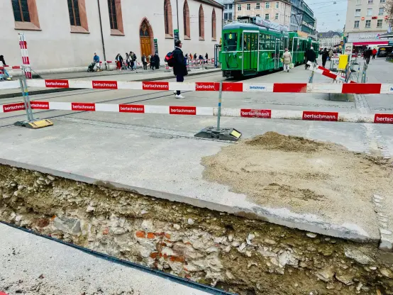 foto von Claraplatz, Basel, mit im hintergrund kirche, tram, und menschen. Im vordergrund eine loch gegraben für neue Leitungen. Alte Steinen unter dem Strasseoberflach sind sichtbar.