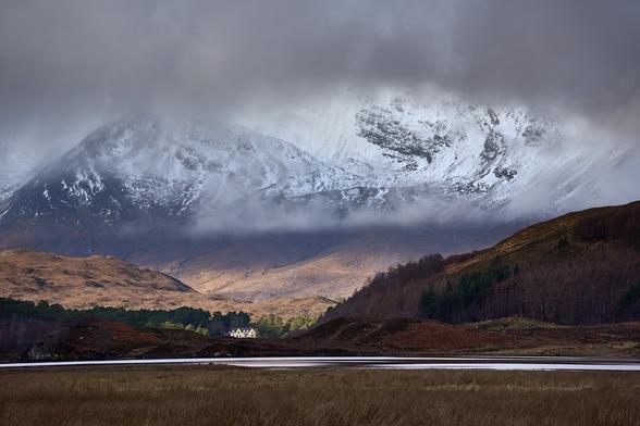 A scene of light playing across a rolling landscape leading to a snowy mountain in the background. Low cloud partially crosses in front of the mountain.