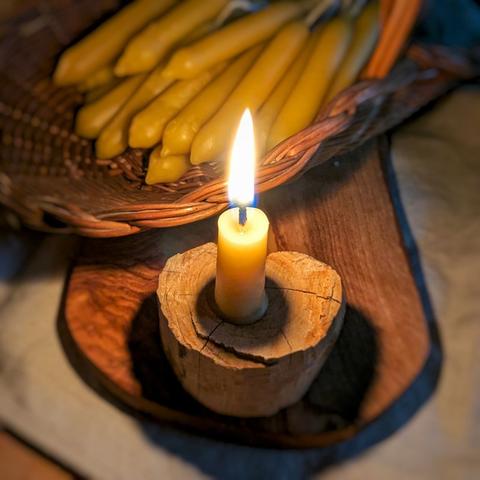 Table with linen cloth. In the background a wicker basket with hand-pulled beeswax candles. In focus is a wooden board with a debarked piece of wood on it, which serves as a candle holder for a burning beeswax candle.