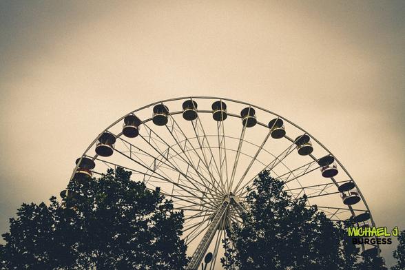 A Ferris wheel rises above the treetops against a muted, vintage-toned sky. The gondolas' subtle colours add a nostalgic charm to the structure's geometric elegance, creating a serene and timeless atmosphere.