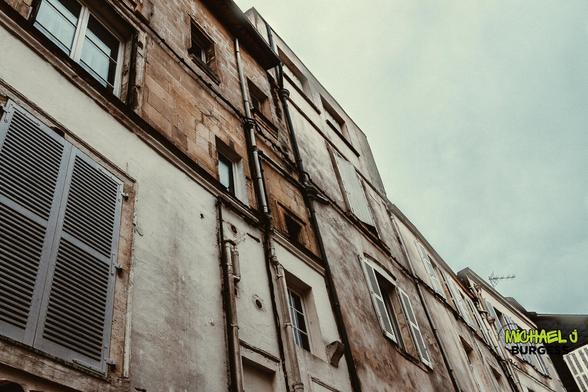 An upward perspective of weathered, historic building facades with worn shutters and exposed pipes set against a muted sky. The texture and age of the structures evoke a sense of timelessness.