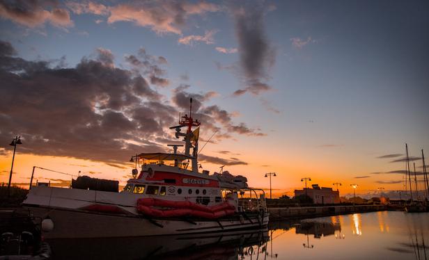 bild im querformat:
im fordergrund das rettungsschiff SeaEye5 am pier. im hinterrund geht die sonne auf und hüllt das ganze bild in einem gold-orangenem licht
