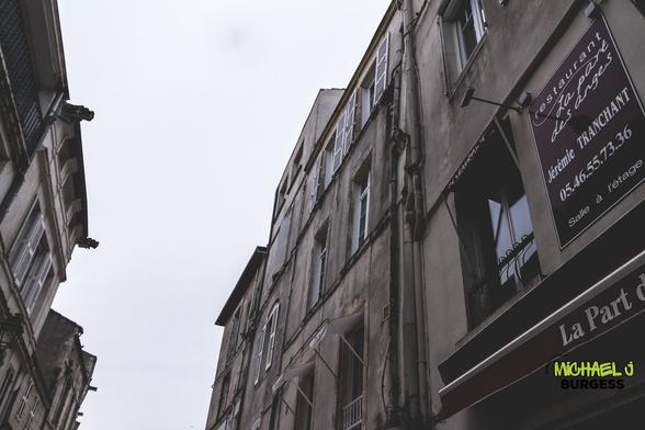 A cloudy urban street scene features old stone buildings with weathered facades and shuttered windows. A restaurant sign for "La Part des Anges" adds local charm, while the overcast sky enhances the subdued, moody atmosphere.