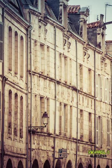 A historic stone facade in La Rochelle featuring elegant arched windows, intricate detailing, and a vintage street lamp. The architectural rhythm and warm tones exude a classic French atmosphere.