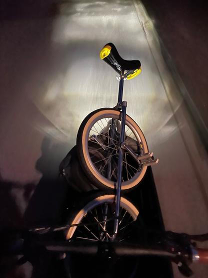 Ein Lastenrad und ein Lichtkegel im Dunkeln. Auf der Ladefläche ist eine Twice, ein Hocheinrad mit zwei übereinander liegenden Rädern.