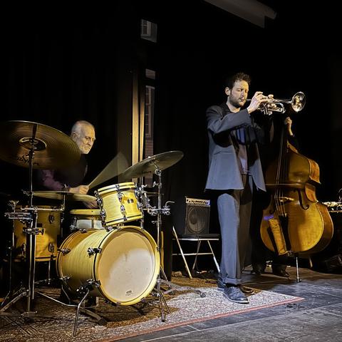 From left to right: Eddie Prévost behind his drum kit, drumsticks ablur in the low light reaching the camera; Silvan Schmid, besuited with trumpet raised to his lips, eyes closed in the heat of the moment; and Tom Wheatley, partly obscured by Silvan, we only see his feet and legs behind his double bass, and a hand on the strings, his face in shadow.
