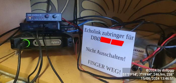 Ganz links unten steht ein Hytera-Funkgerät. Auf dem Funkgerät ein kleiner Mini-PC. Da drüber ein "USB Interface III". 

Auf der rechten Seite das Netzteil, davor ein Laminiertes Schild mit
"Echolink zubringer für 
DB0[Zensiert] / [Zensierter Ortsverband]

Nicht Ausschalten!

FINGER WEG!
"