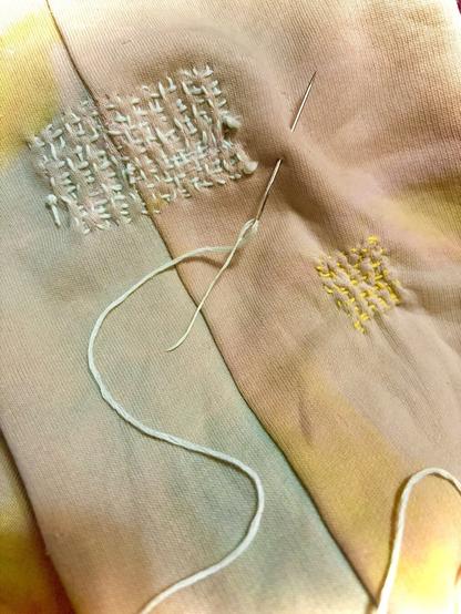 Close up photo of tie-dye yellow, teal and pink colored fabric with two sections of cross hatched patch work using embroidery thread in yellow and teal color with the needle pined to the cloth with remaining teal thread through the eye of the needle