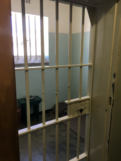 A photo of Nelson Mandela‘s prison cell on Robben Island. There is a window with bars in the background and the entrance doorway is also metal bars the 7‘ x 9‘ room has a small table visible.