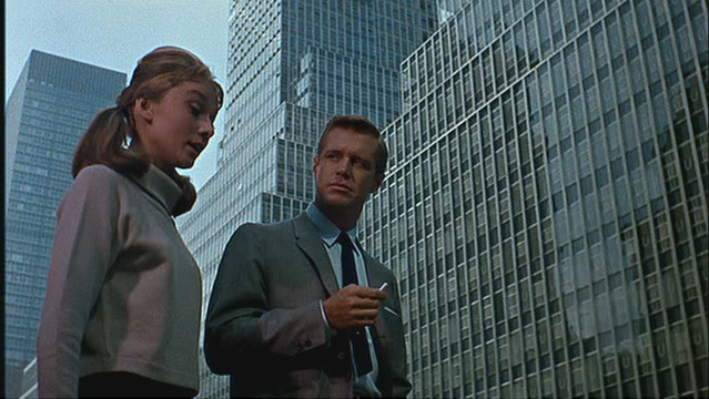 Fotograma de la película "Breakfast at Tiffany's" 💎 (1963) de Blake Edwards. Una mujer con el pelo recogido en dos coletas y un jersey de cuello alto gris claro está de pie junto a un hombre en traje gris que sostiene un cigarrillo. Ambos miran hacia arriba a los imponentes rascacielos de cristal y acero que los rodean en una ciudad moderna.  El fondo está dominado por la arquitectura moderna de la ciudad.