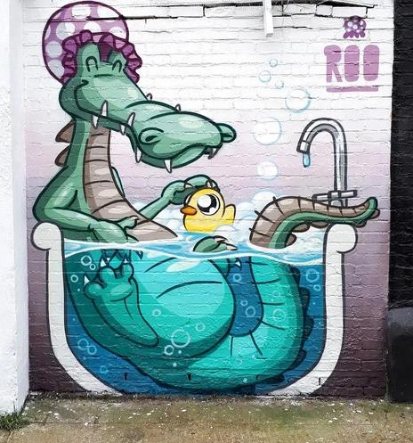 Streetartwall. A mural with a funny crocodile in a bathtub was sprayed /painted on a white brick wall. The green crocodile lies smiling in a transparent bathtub wearing a purple shower cap. The bathtub is a little too small, but there is still room for his yellow squeaky duck.