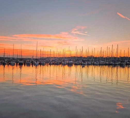 Pastel sunrise over a marina lined with sailboats. Bright colors swirl above the horizon as sunrise approaches,  casting shades of pink, yellow, and orange across a light blue sky, bright enough to also make reflections upon still river waters below. The horizon is slightly obscured by a long row of sailboats tied up in a marina, also casting their reflection upon the water below.
