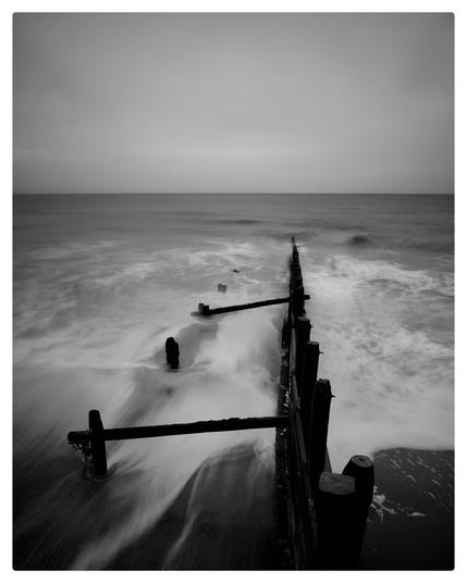 Black and white image of groynes heading out to sea. The sky is flat grey and the sea swirls around the wooden posts making patterns and shapes