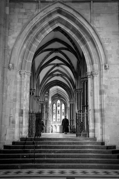 Taken inside a cathedral looking up stone stairs, through a pointed arch, into a chapel. The chapel has a high vaulted ceiling supported by pillars and beams. There is a stained glass window at the far end. A figure is walking through the chapel, with their back to camera.