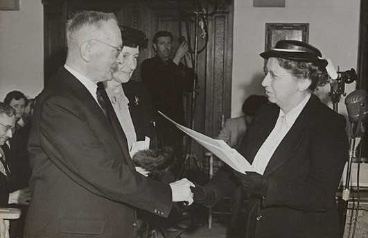 Anna de Waal shaking hands with someone. She is a white woman in a dark suit and hat.