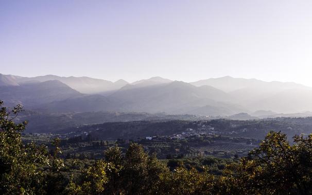 A hazy mountain landscape in Crete, Greece, with layers of rugged peaks fading into the soft evening light. Below, a valley dotted with villages and olive groves showcases the island’s diverse topography.