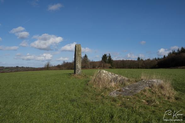 Photo en couleur, présentant trois menhirs dont deux sont couchés au milieu d'un champ d'herbe sous un ciel pratiquement sans nuage. Le champ se situe au sommet d'une colline et en arrière-plan, on voit d'autres sommets, pratiquement de la même altitude, mais couverts de forêt. La photo a été prise pratiquement depuis le sol en contre-plongée avec les deux pierres couchées au premier plan sur la gauche, alors que la pierre toujours debout se situe au milieu du cadrage sur un second plan.