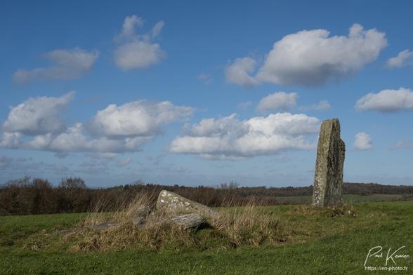 Photo en couleur, présentant trois menhirs dont deux sont couchés au milieu d'un champ d'herbe sous un ciel pratiquement sans nuage. Le chant se situe au sommet d'une colline et en arrière-plan, on voit d'autres sommets, pratiquement de la même altitude, mais couverts de forêt. La photo a été prise, pratiquement depuis le sol en contre-plongée avec les deux pierres couchées au premier plan au centre de la photo, alors que la pierre toujours debout, se trouve dans un second plan sur la droite.