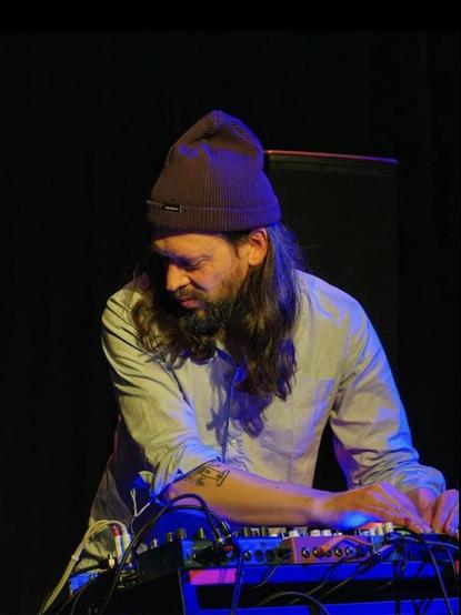 Ach Zukunft, a man with a beard, long dark hair and brown beanie at electronic devices to make experimental sound or visuals. He wears a white shirt.