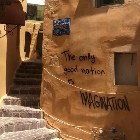 A piece of street art graffiti on a warm-toned ochre wall in Chania, Crete, Greece. The message, written in black paint, reads: "The only good nation is IMAGINATION," with "IMAGINATION" emphasized in large, bold letters. Above the graffiti, a blue and white street sign marks "Douka Dimitriou" street. The surrounding scene features stone steps leading up to a picturesque, sunlit alleyway with Mediterranean architectural details, including arched doorways and wrought-iron elements. The graffiti blends seamlessly with the rustic charm of the historic setting, creating a thought-provoking contrast.