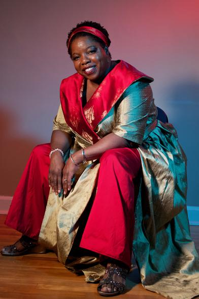 A black woman sits 3/4 facing the camera , leaning forward comfortably, wearing a gold and red silk wrap dress over red loose cut pants. She has a matching red and gold silk head band.