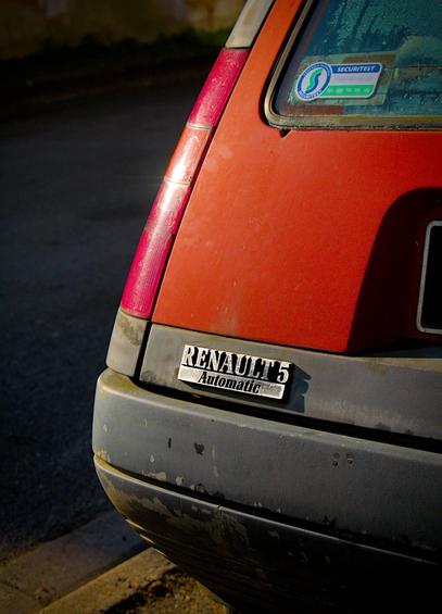 La photo montre l'arrière d'une voiture, en l'occurrence une Renault 5. Elle porte un badge « Automatic » et un autocollant est apposé sur la vitre arrière. La voiture est de couleur rouge.