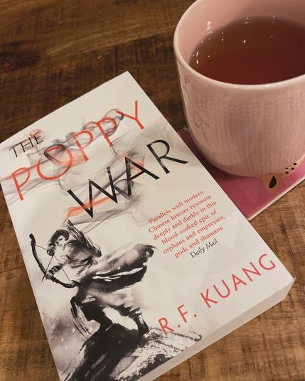 A paperback copy of The Poppy War by R.F. Kuang and a pink mug of tea on a wooden table.