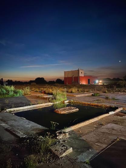 Scenery of a sunset at a industrial grounds - old chemical plant. 

In the foreground; a dark puddle of water in the remains of a building foundation. Wild grass and plants sprouting out through the asphalt and concrete ground.

Hard light source from the side from flood lights giving a high contrast fore ground. 

Background is of shipping container buildings and gradual sunset colours. Some night stars can be seen.