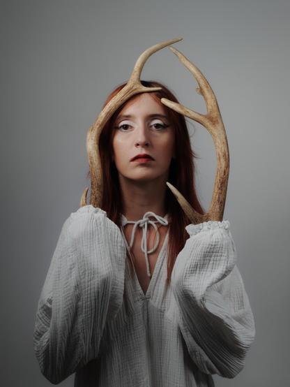 The image shows a person with red hair holding antlers in front of their face. They are wearing a white blouse with long sleeves. The background is plain and gray.