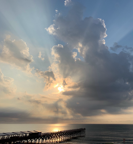 Sun rays shining through clouds that are breaking up after a storm out over the ocean; the sun is reflected onto the calm dark gray-blue water; there is a wooden pier at the bottom left of the image.