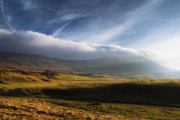 A dramatic landscape near Chapel-le-Dale, with Ingleborough mountain dominating the scene. The foreground features rolling hills covered in lush green grass, interspersed with patches of brown heather. A winding path cuts through the landscape, leading towards the base of the mountain. The sky is a striking mix of deep blue and soft white clouds, with a thick blanket of cloud resting on the summit of Ingleborough, creating a moody atmosphere. The light casts gentle shadows across the terrain, enhancing the contours of the hills and adding depth to the scene.