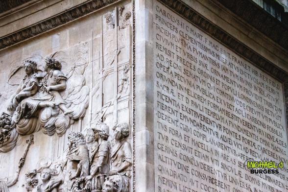 This is a close-up of an intricately carved stone relief on a historic London monument. The relief features classical figures and detailed Latin inscriptions. It portrays a regal scene with allegorical figures, symbolizing history and legacy.