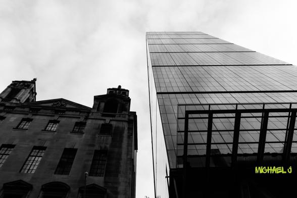 This dramatic black-and-white shot of London's skyline contrasts a historic classical building and a sleek modern glass skyscraper. The low-angle perspective emphasizes the city's architectural evolution, with reflections and sharp lines dominating the frame.