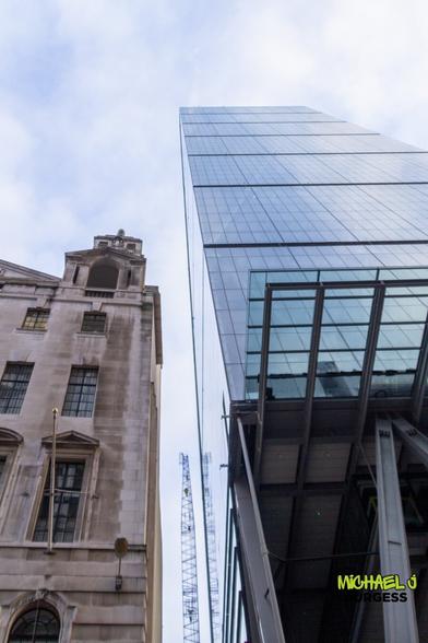 This striking juxtaposition of old and new architecture in London features a historic stone building alongside a towering modern glass skyscraper. The skyscraper's reflective surfaces contrast with the older structure's aged texture, symbolizing the city's blend of heritage and innovation. The construction crane in the background hints at continuous urban development.