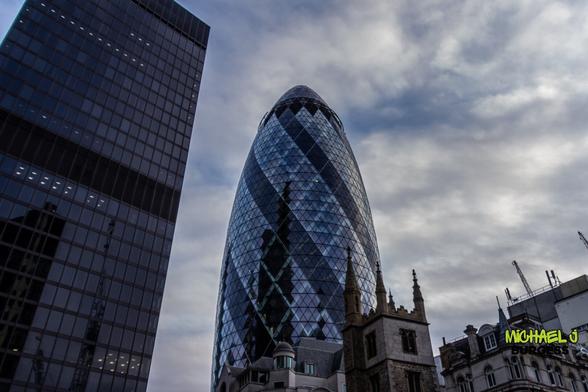 This is a striking view of London's iconic Gherkin skyscraper, its curved glass facade reflecting the cloudy sky. The modern architecture contrasts sharply with the surrounding historic buildings, blending the city's rich past with its cutting-edge present. The dark tones emphasize the dramatic interplay of light and reflections.