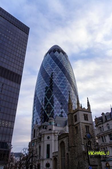A dynamic cityscape featuring London's Gherkin, its reflective glass façade towering over a historic stone church. The blend of modern and old architecture creates a compelling contrast, showcasing the city's architectural evolution under a soft blue sky.