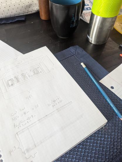 Graph paper notebook with a light box sketch
A box with the word charlie in 3 dimensions, with measurements. A mug and a stainless tumber of coffee in the background of a messy dining table.